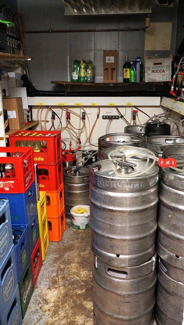 Lagerraum mit gestapelten Bierfässern und Getränkekisten, umgeben von Versorgungsleitungen.
