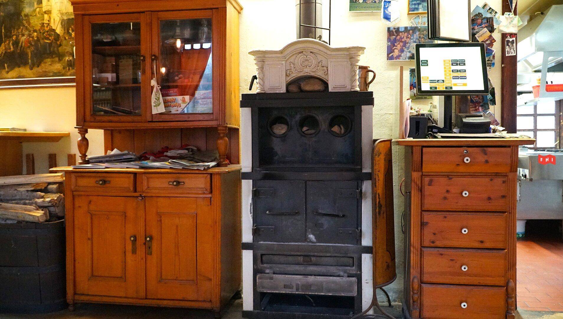 Traditioneller Kachelofen neben einem antiken Holzschrank und einer Kommode, mit einem modernen Monitor.