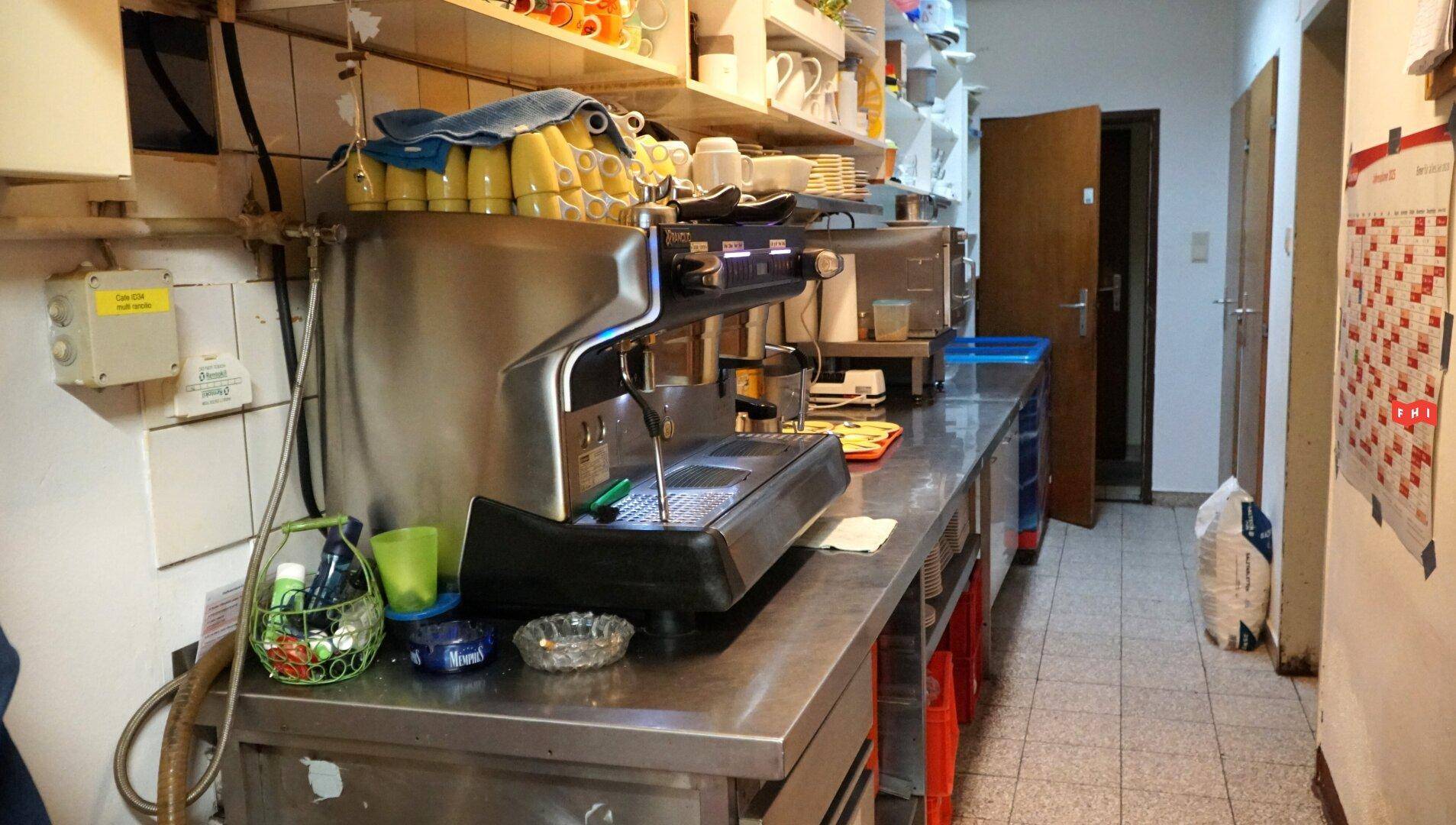 Schmaler Arbeitsbereich mit professioneller Kaffeemaschine, Edelstahlarbeitsplatte und Regalen voller Tassen.