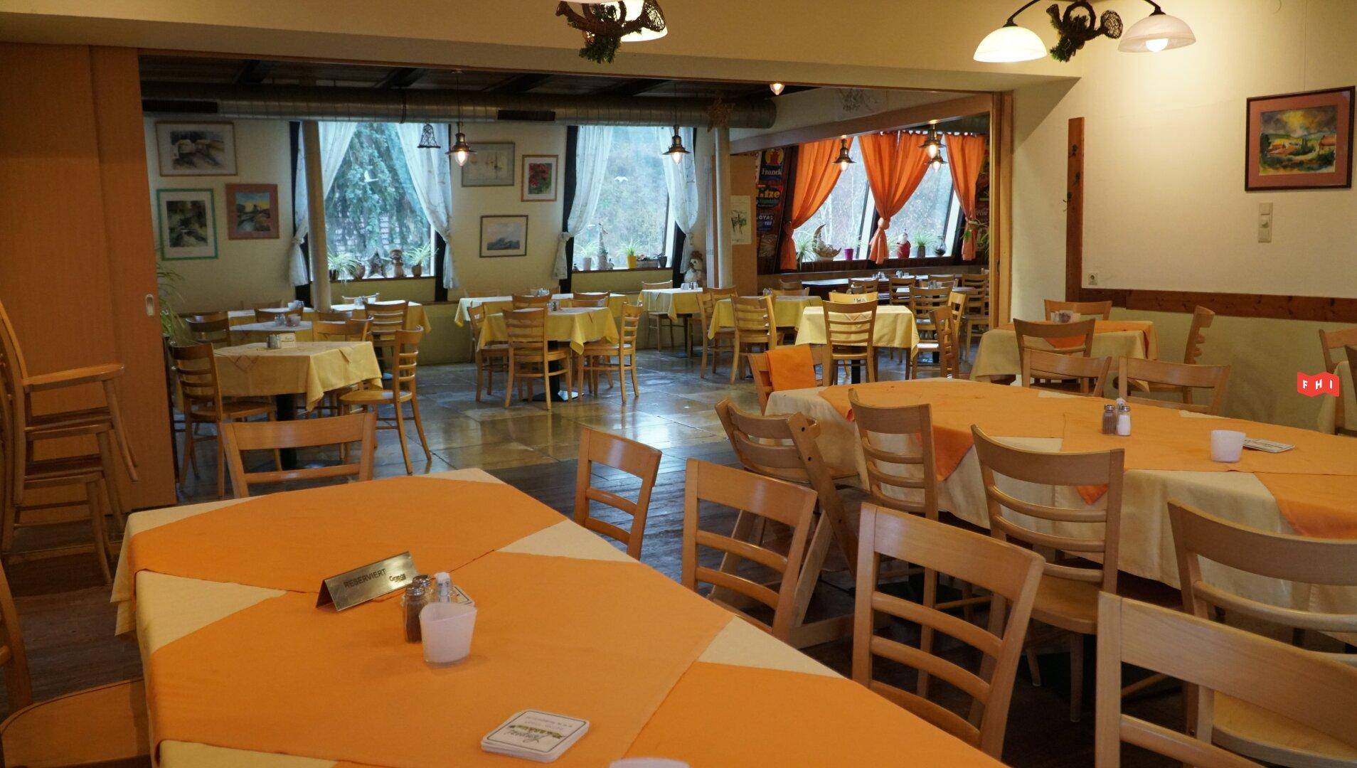 Großer Speisesaal mit vielen Tischen, orangefarbenen Tischdecken und Fenstern, die viel Tageslicht hereinlassen.