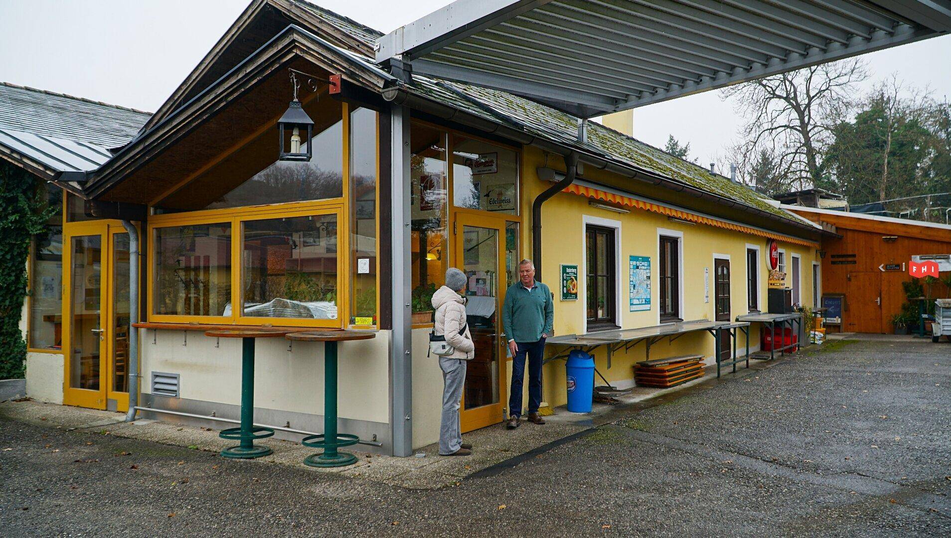 Gelbes Gebäude mit Verkaufsbereich im Freien und zwei Personen davor, umgeben von einer ländlichen Umgebung.