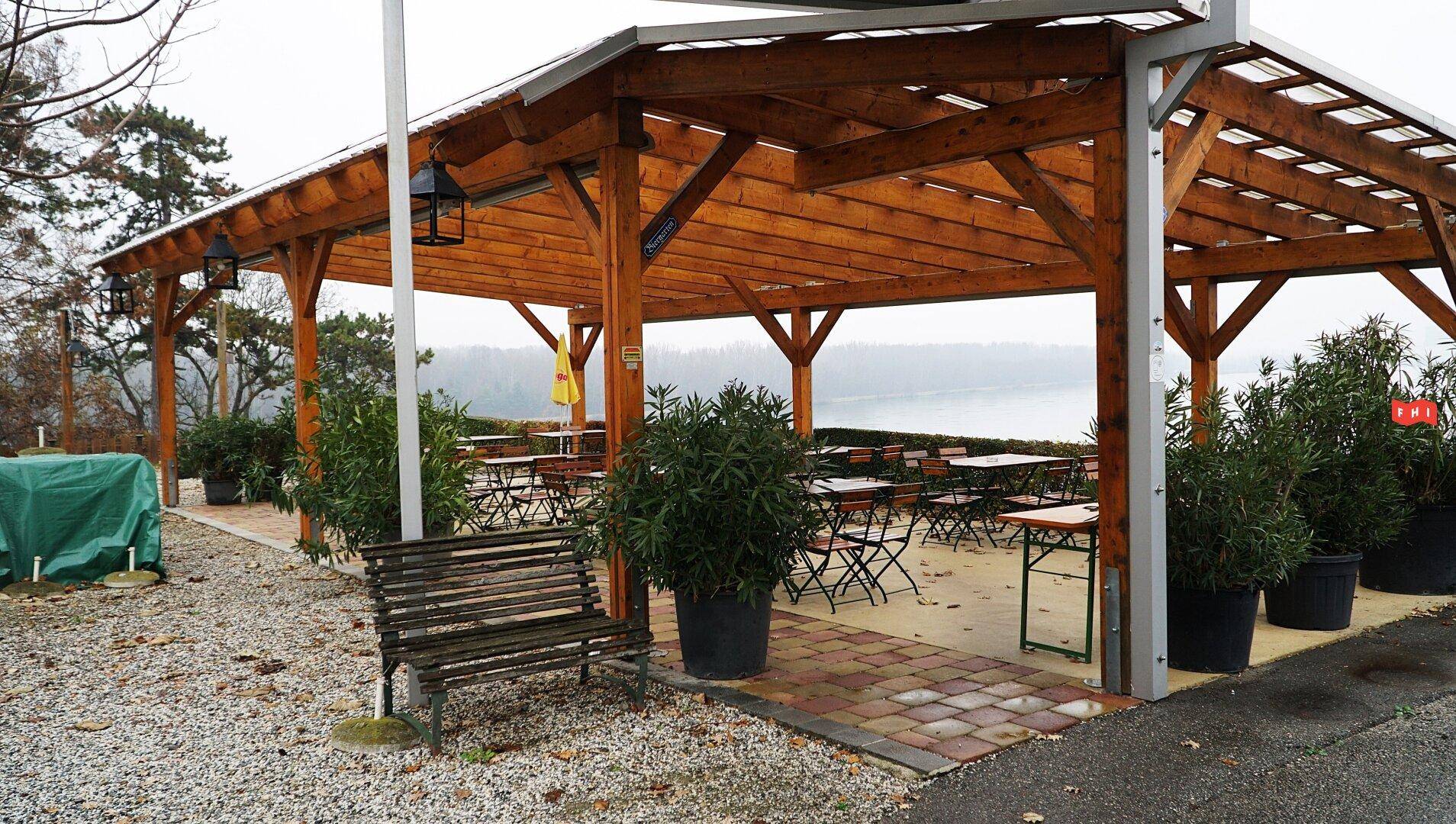 Überdachte Terrasse mit Holzdach, Sitzbänken und Tischen, die einen weiten Blick auf den See bietet.