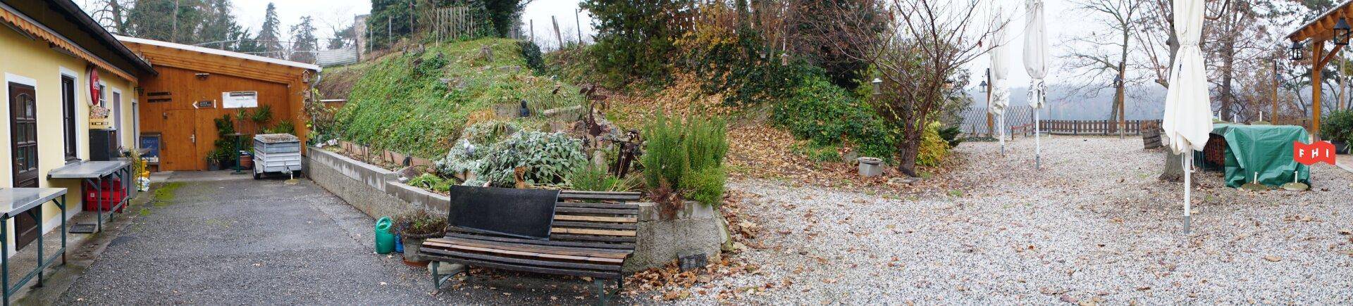 Weitläufiger Gartenbereich mit einem Gebäude links, einem grünen Hang und einer überdachten Terrasse.