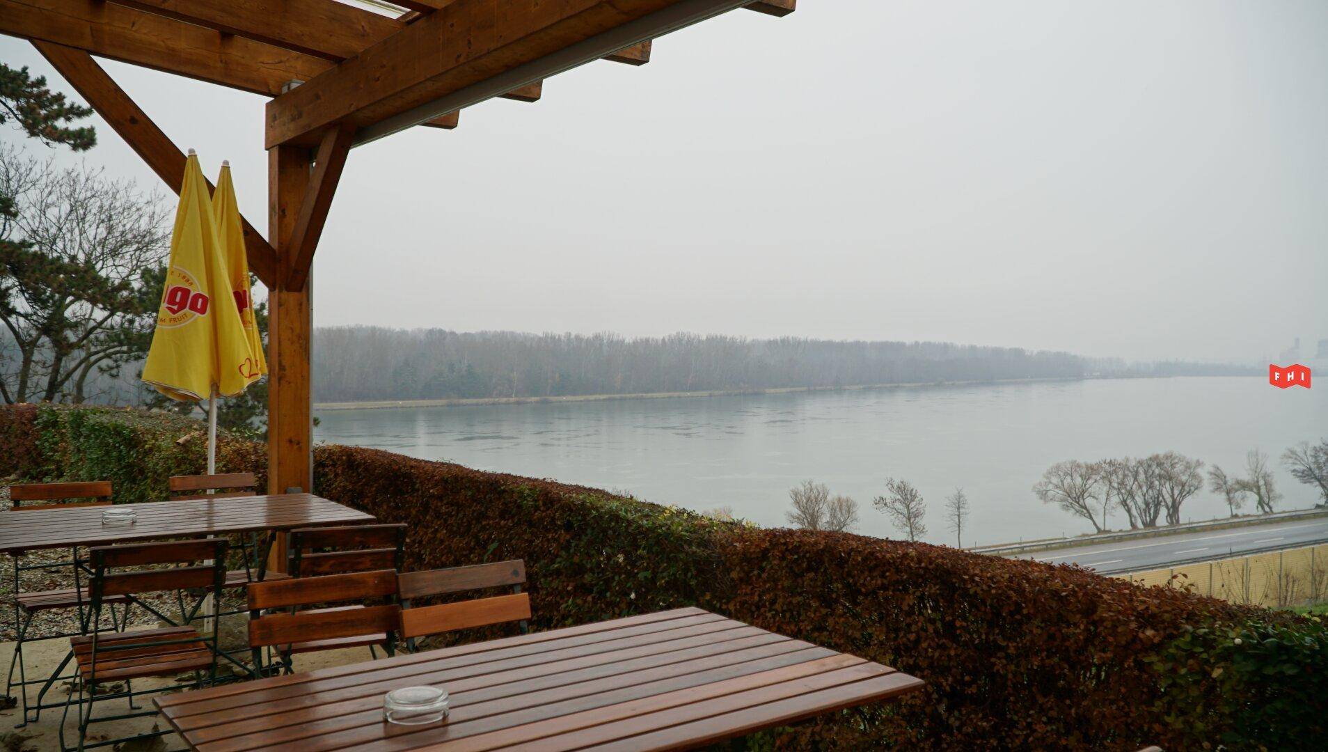 Überdachte Terrasse mit Tischen und Stühlen, die einen malerischen Blick auf den Fluss oder See bietet.
