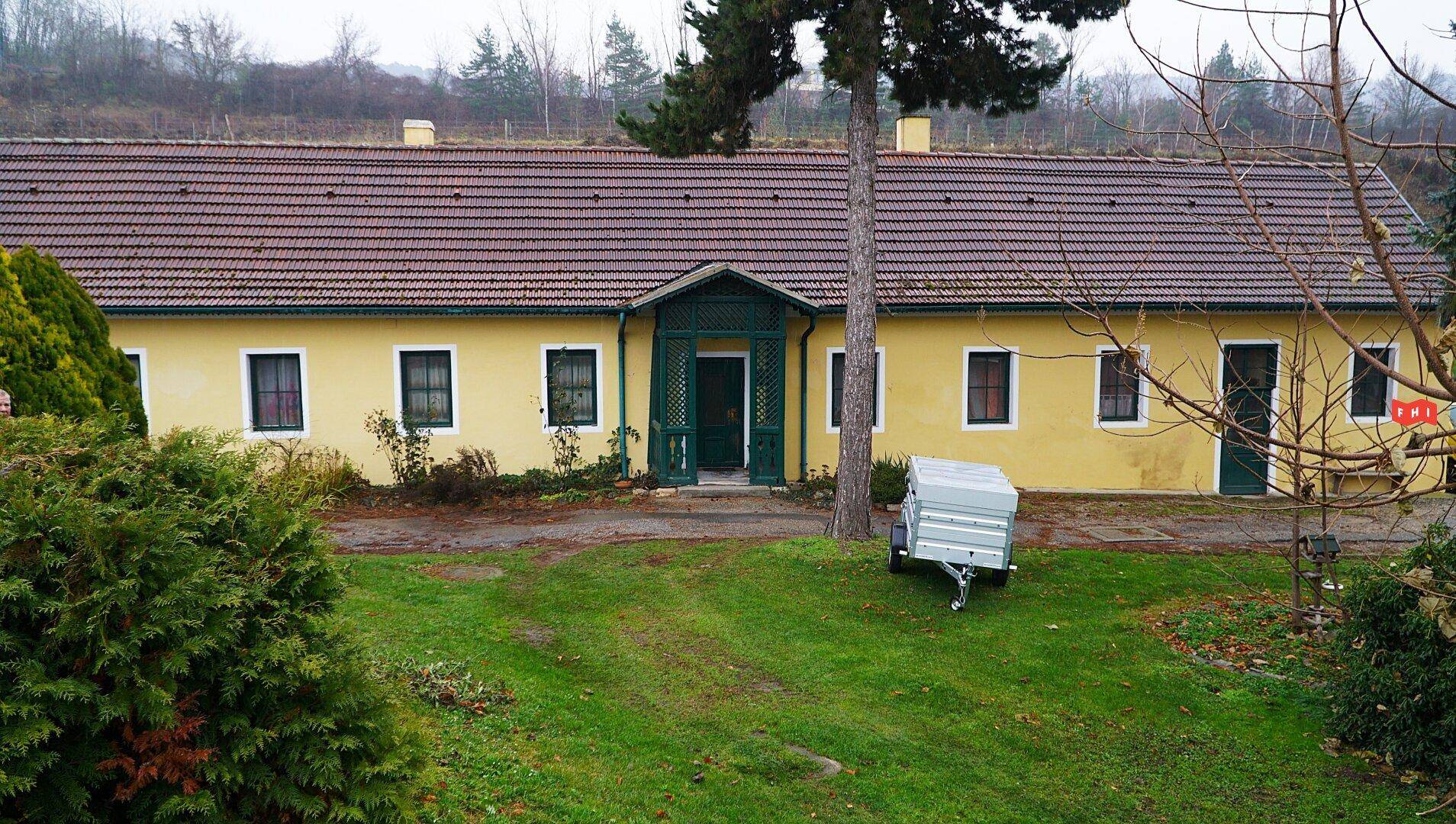 Gelbes Gebäude mit Ziegeldach und grünen Fensterläden, umgeben von einem gepflegten Rasen und Bäumen.