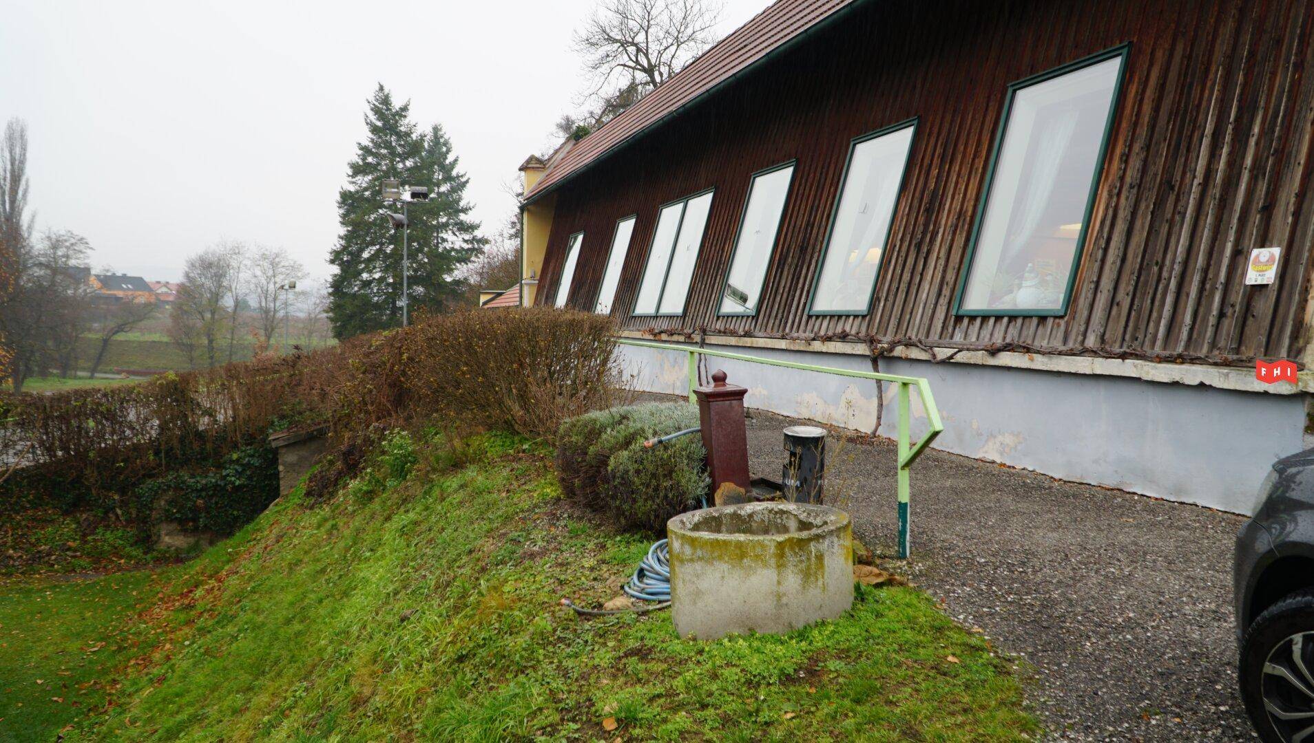 Gebäude mit Holzfassade und schrägen Fenstern, umgeben von einem grünen Hang und einem alten Brunnen.