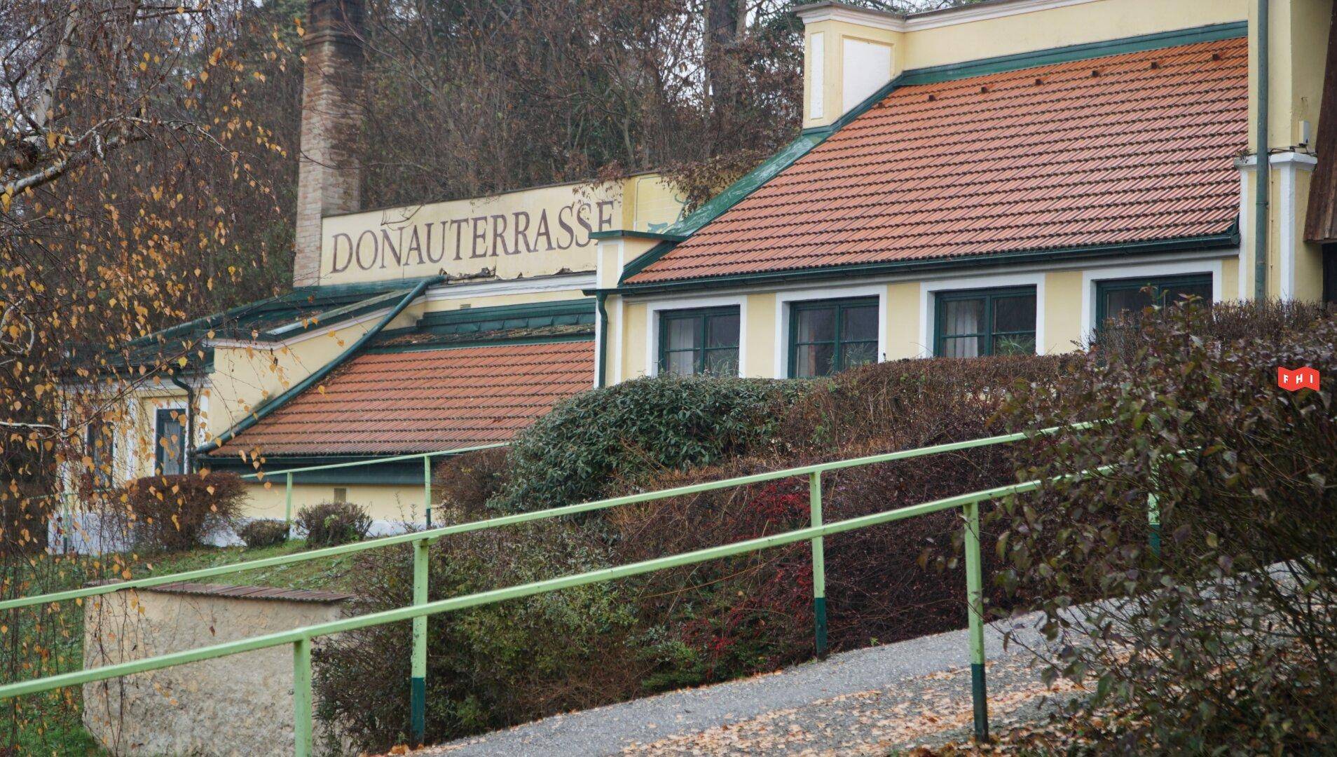 Gebäude mit dem Schriftzug "DONAUTERRASSE", gelber Fassade und Ziegeldach, mit einer grünen Rampe im Vordergrund.