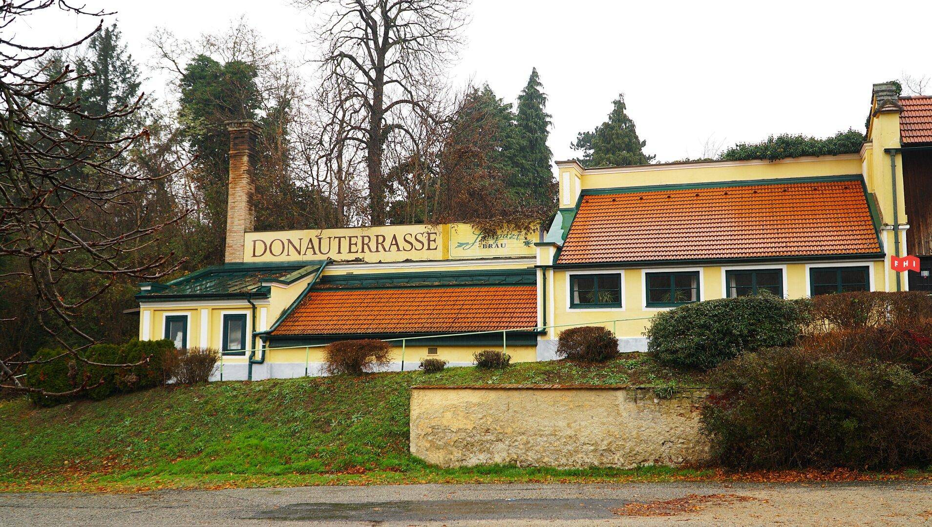 Gebäude mit dem Schriftzug "DONAUTERRASSE", gelber Fassade, Ziegeldach und umgebender Vegetation.
