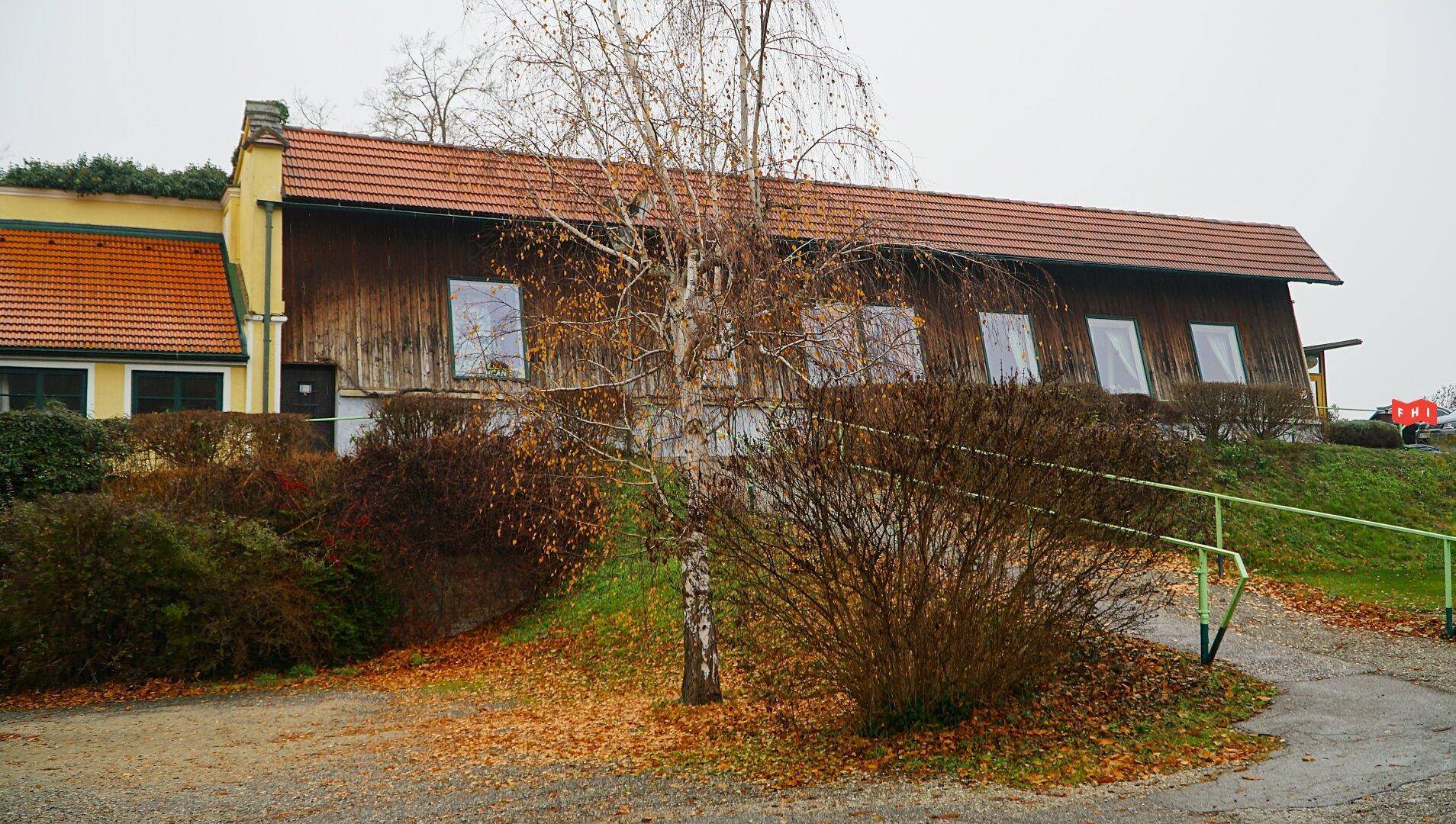 Ländliches Gebäude mit Holzfassade und Ziegeldach, umgeben von Bäumen und Sträuchern im Herbst.