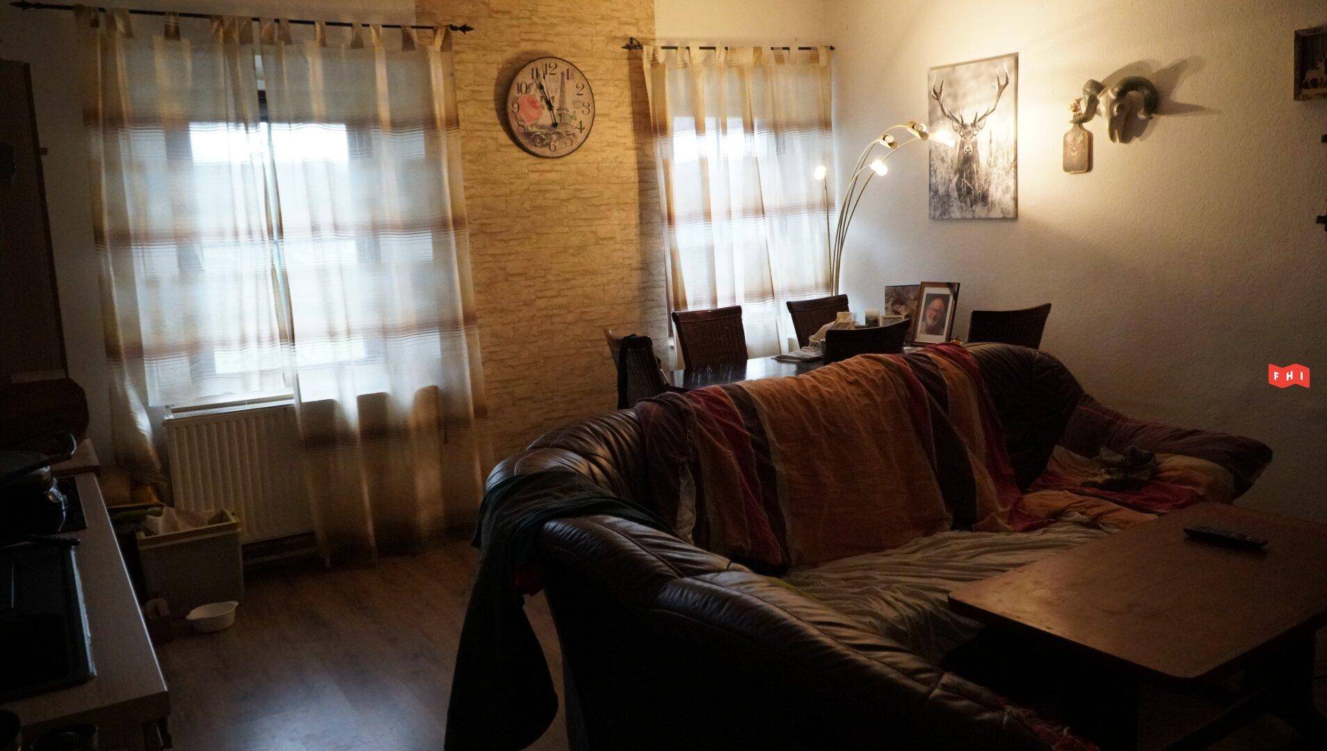 Gemütlicher Wohn-Essbereich mit Ledersofa, Couchtisch und Esstisch, beleuchtet durch Fenster mit Vorhängen.