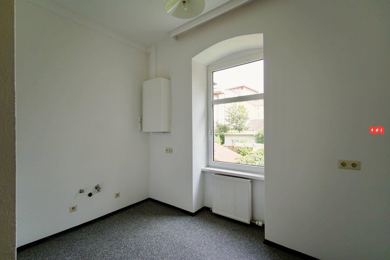 Leerer Raum mit Teppichboden, Fenster und Wandanschlüssen, ideal für eine Küche oder Abstellraum.