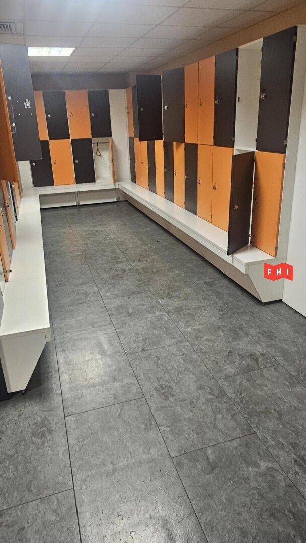 Umkleideraum mit Reihen von orange-braunen Spinden und Sitzbänken auf grauem Fliesenboden.