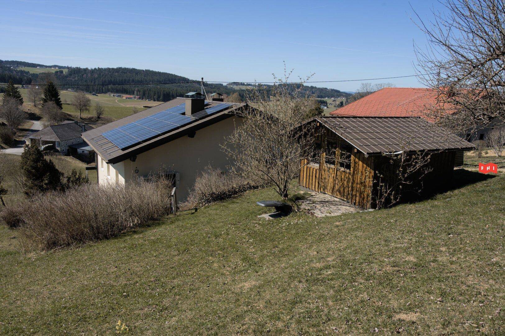 Haus mit Solaranlage auf dem Dach und einem kleinen Holzschuppen in ländlicher Umgebung.