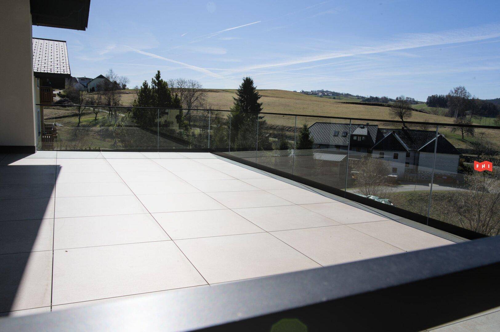 Geräumige, geflieste Terrasse mit Glasgeländer und weitem Blick über die grüne Landschaft.