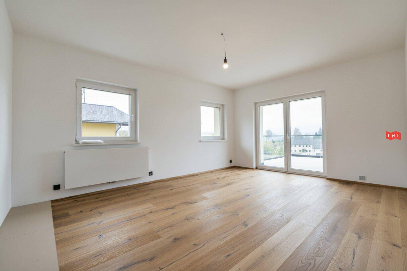 Heller Raum mit Holzboden, großen Fenstern und Zugang zu einem Balkon oder einer Terrasse.