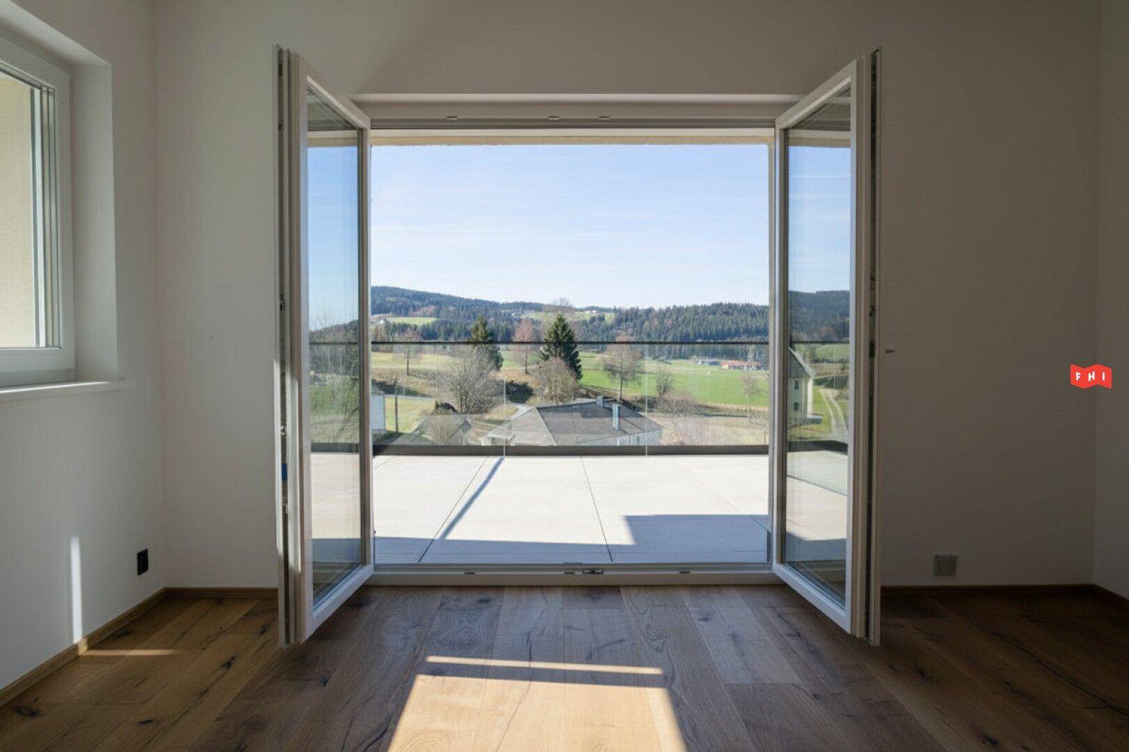 Heller Raum mit Holzboden und großen Fenstertüren, die zu einer Terrasse mit Weitblick führen.