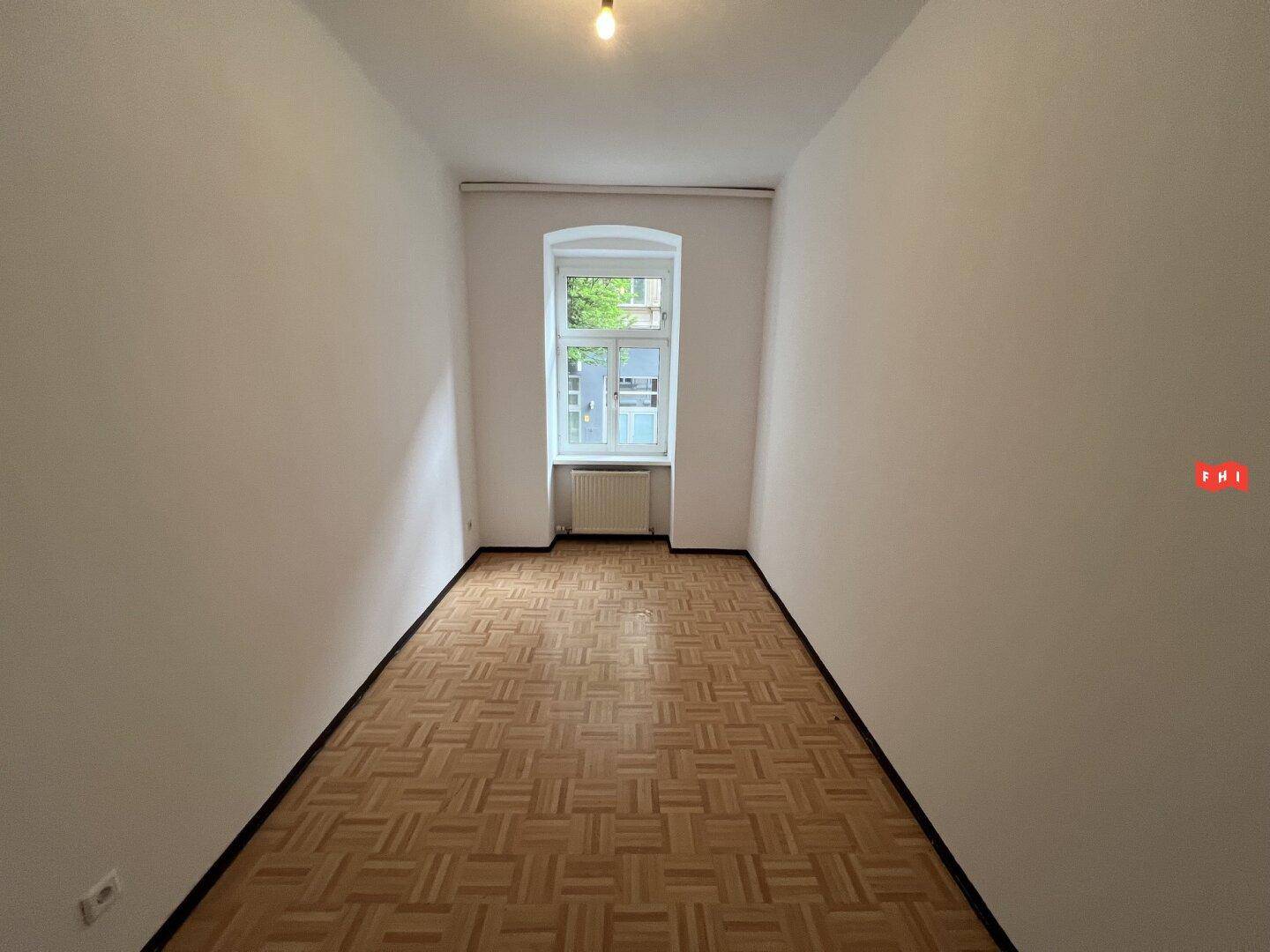 Leerer Raum mit Fischgrätparkettboden und einem Fenster, das den Raum hell erscheinen lässt.