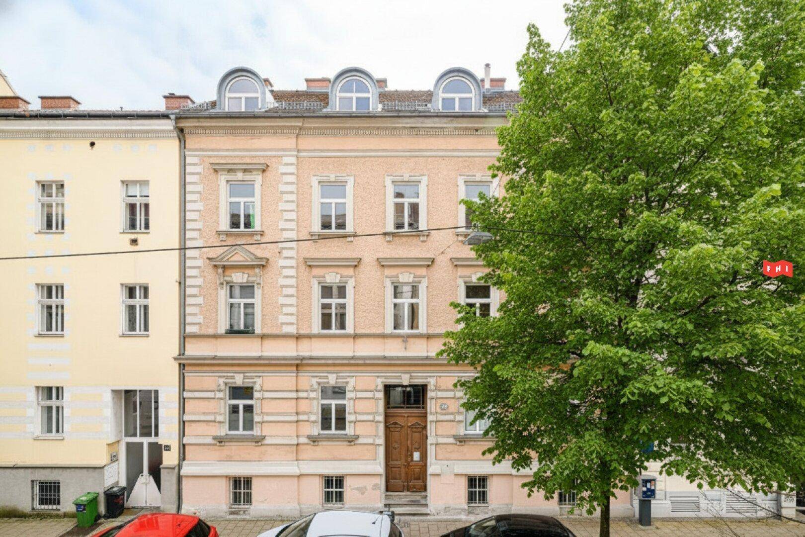 Historische Fassade eines Mehrfamilienhauses mit vielen Fenstern und einem großen Baum davor.
