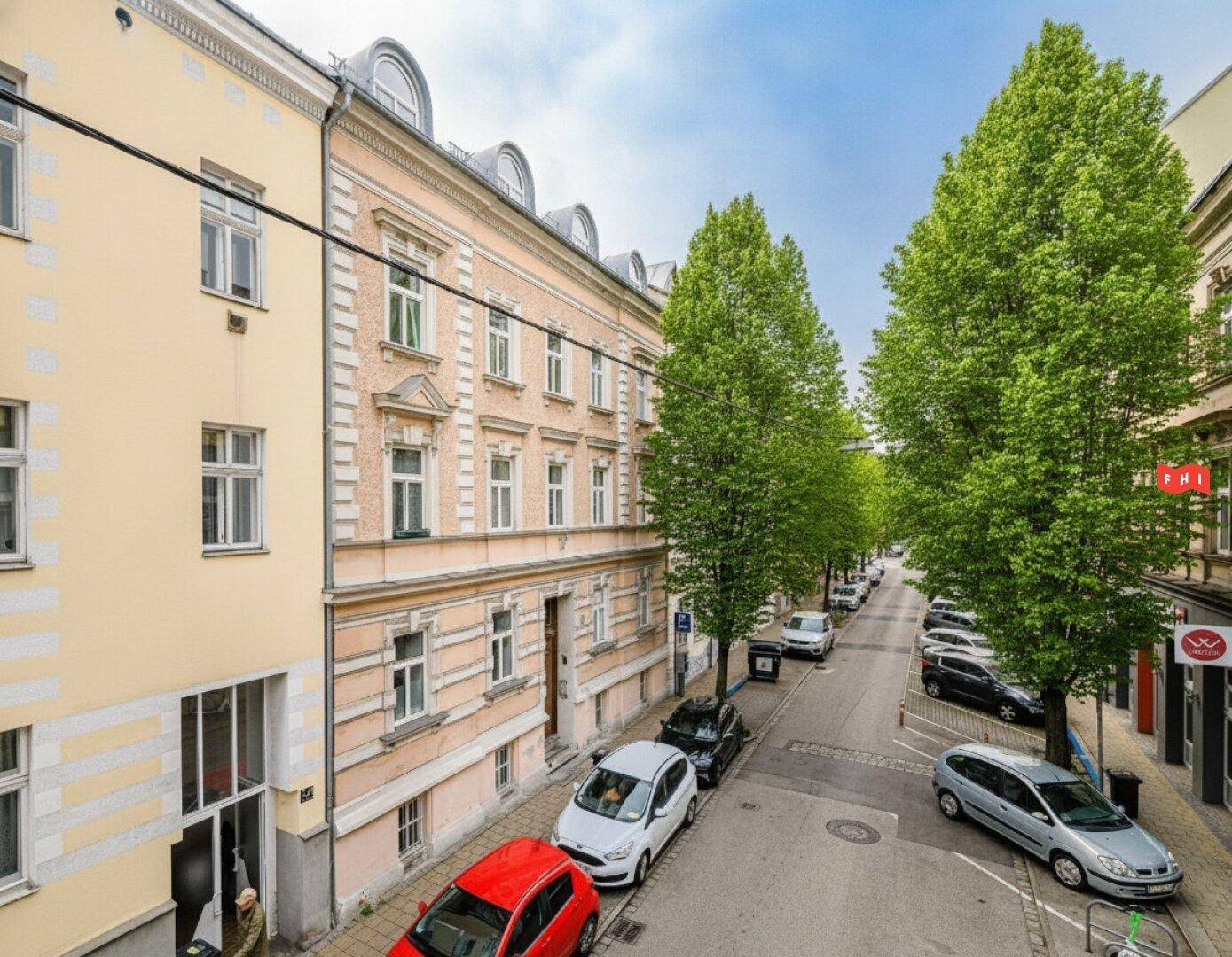 Historische Fassade eines Mehrfamilienhauses an einer belebten Straße mit parkenden Autos und Bäumen.