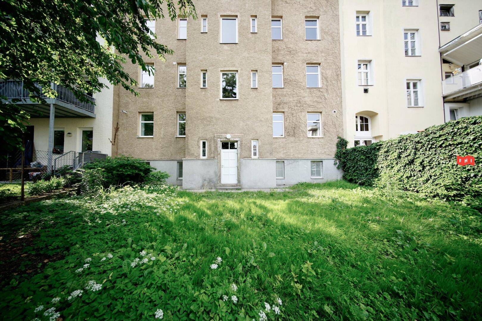 Grüner Hinterhof mit Rasenfläche und einer Gebäudefassade im Hintergrund, umgeben von Pflanzen.