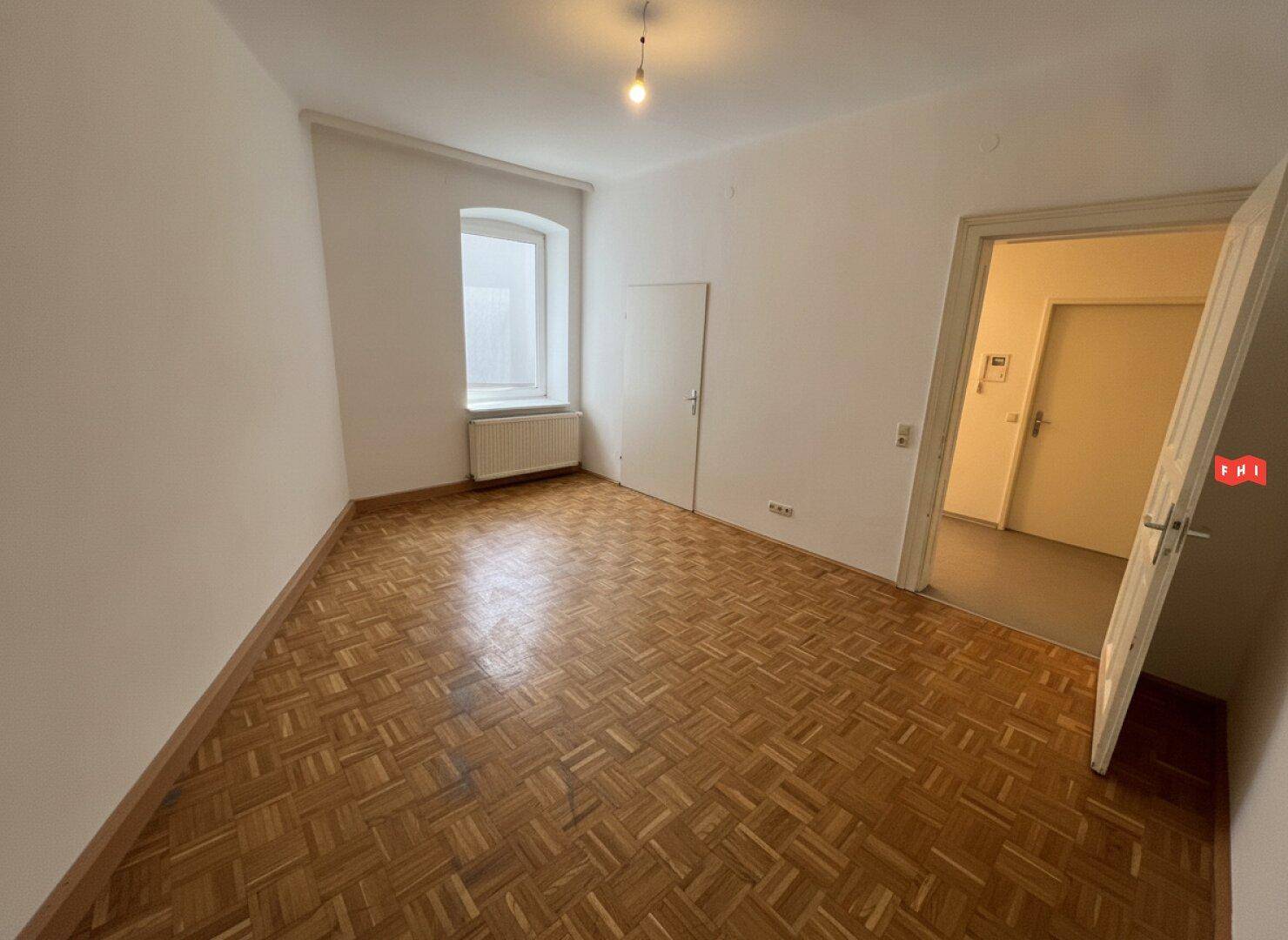 Lichtdurchfluteter Raum mit Parkettboden, einem Fenster und Zugang zu weiteren Zimmern.