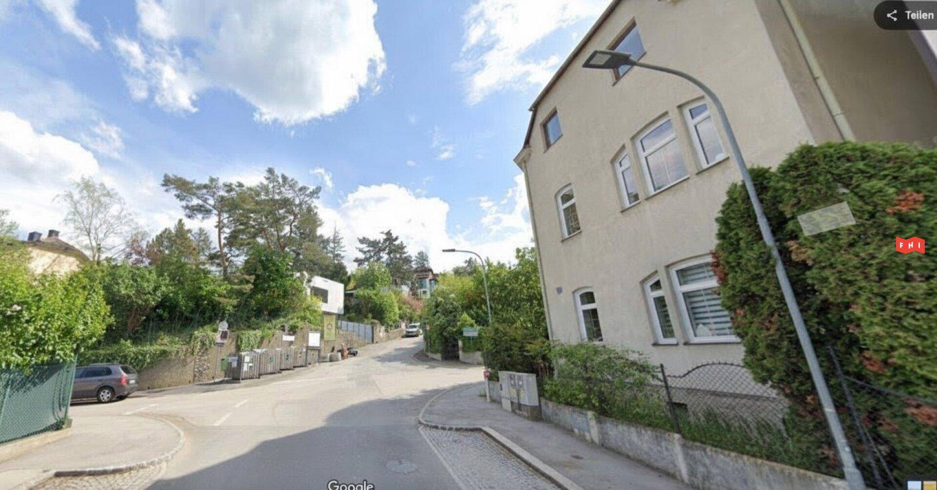 Blick auf die Straße und das Wohngebäude mit viel Grün in der Umgebung unter blauem Himmel.