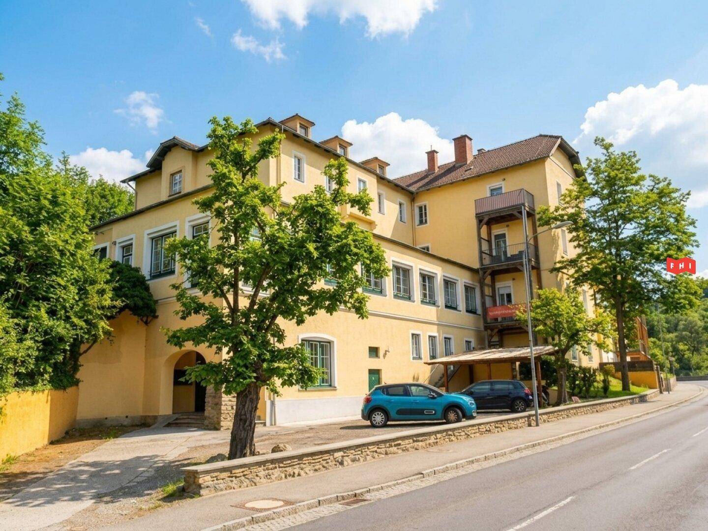 Historisches gelbes Gebäude mit vielen Fenstern und Bäumen, gelegen an einer Straße.