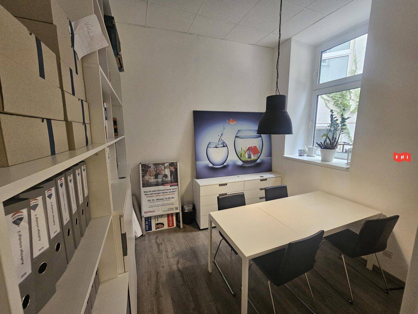 Kleineres Bürozimmer mit Besprechungstisch, Regalen und einem Fenster für Tageslicht.