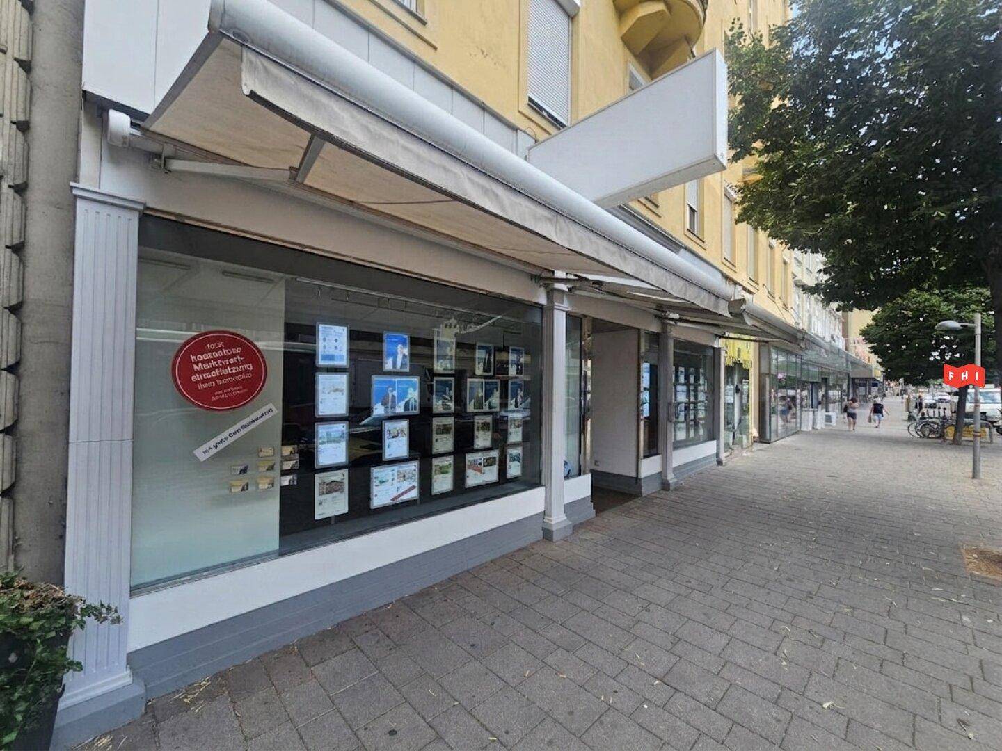 Seitenansicht eines Immobilienbüros mit Schaufenstern und Markisen entlang einer städtischen Geschäftsstraße.