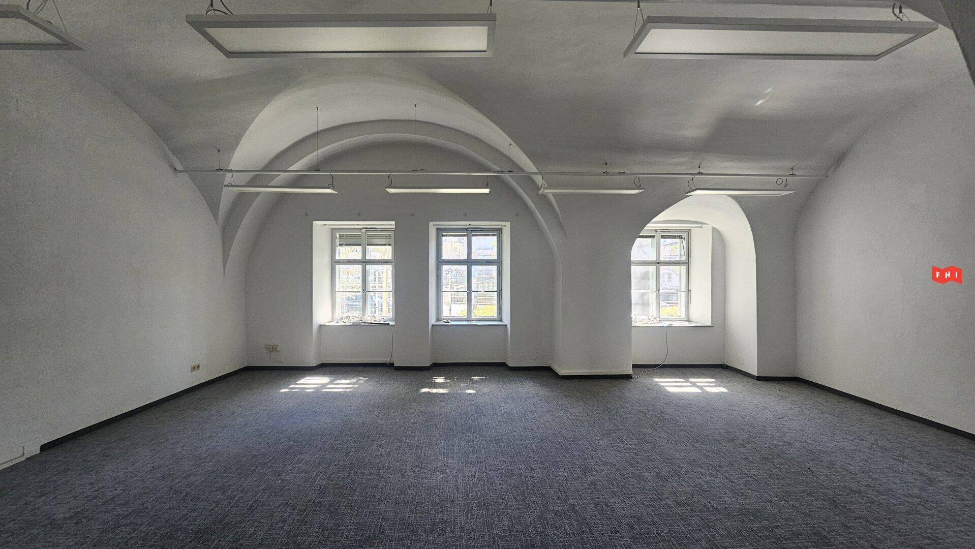 Geräumiger Büroraum mit weißer Gewölbedecke, Teppichboden und großen Fenstern, die viel Licht hereinlassen.