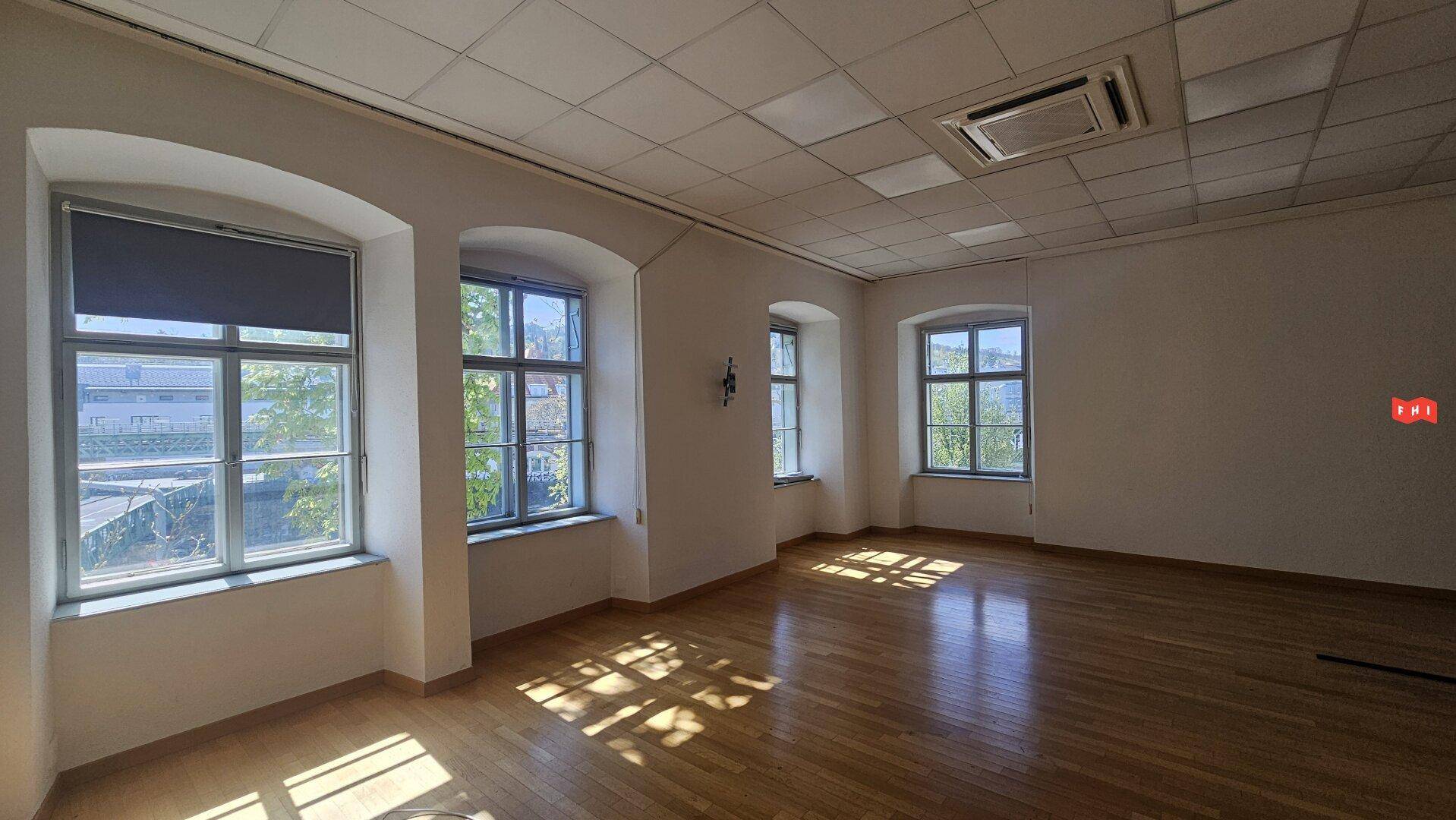 Heller Büroraum mit Holzparkettboden, abgehängter Decke und großen Fenstern, die Tageslicht spenden.