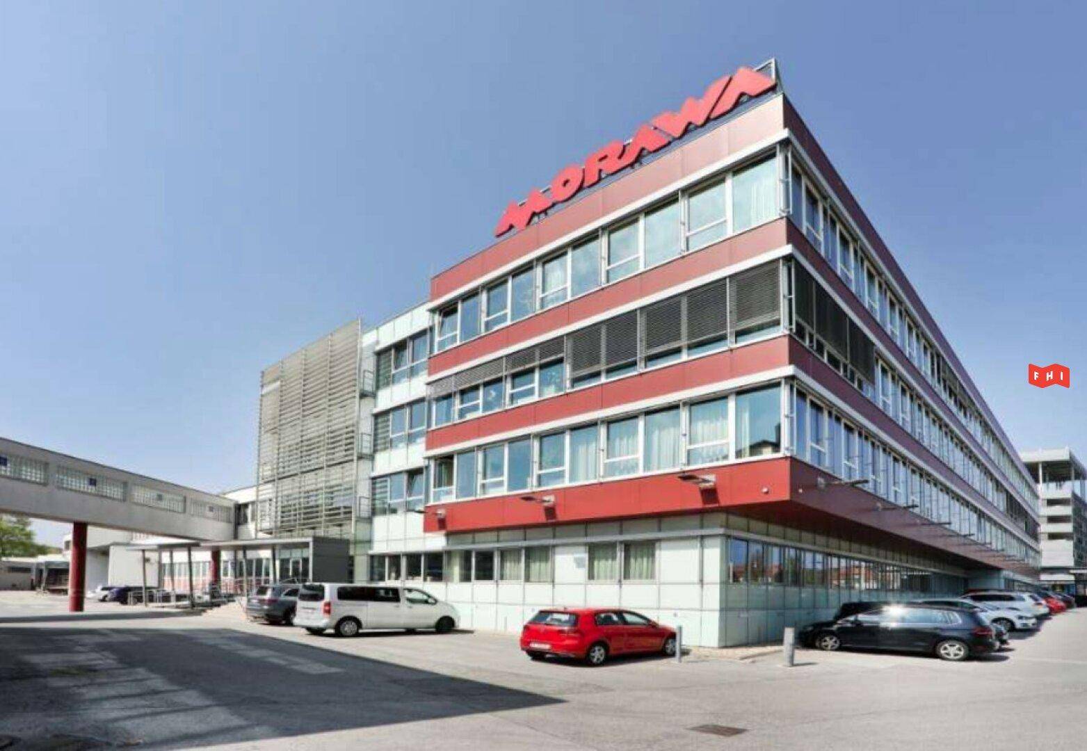 Modernes Bürogebäude mit roter Fassade, großen Glasflächen und einem Parkplatz davor.