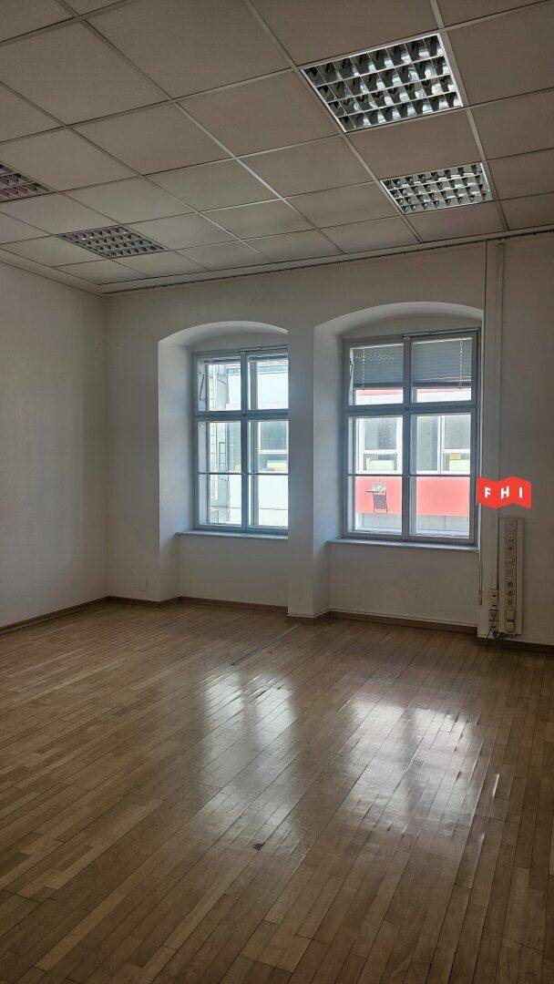 Heller Büroraum mit Holzparkettboden, abgehängter Decke und zwei großen Fenstern mit Jalousien.