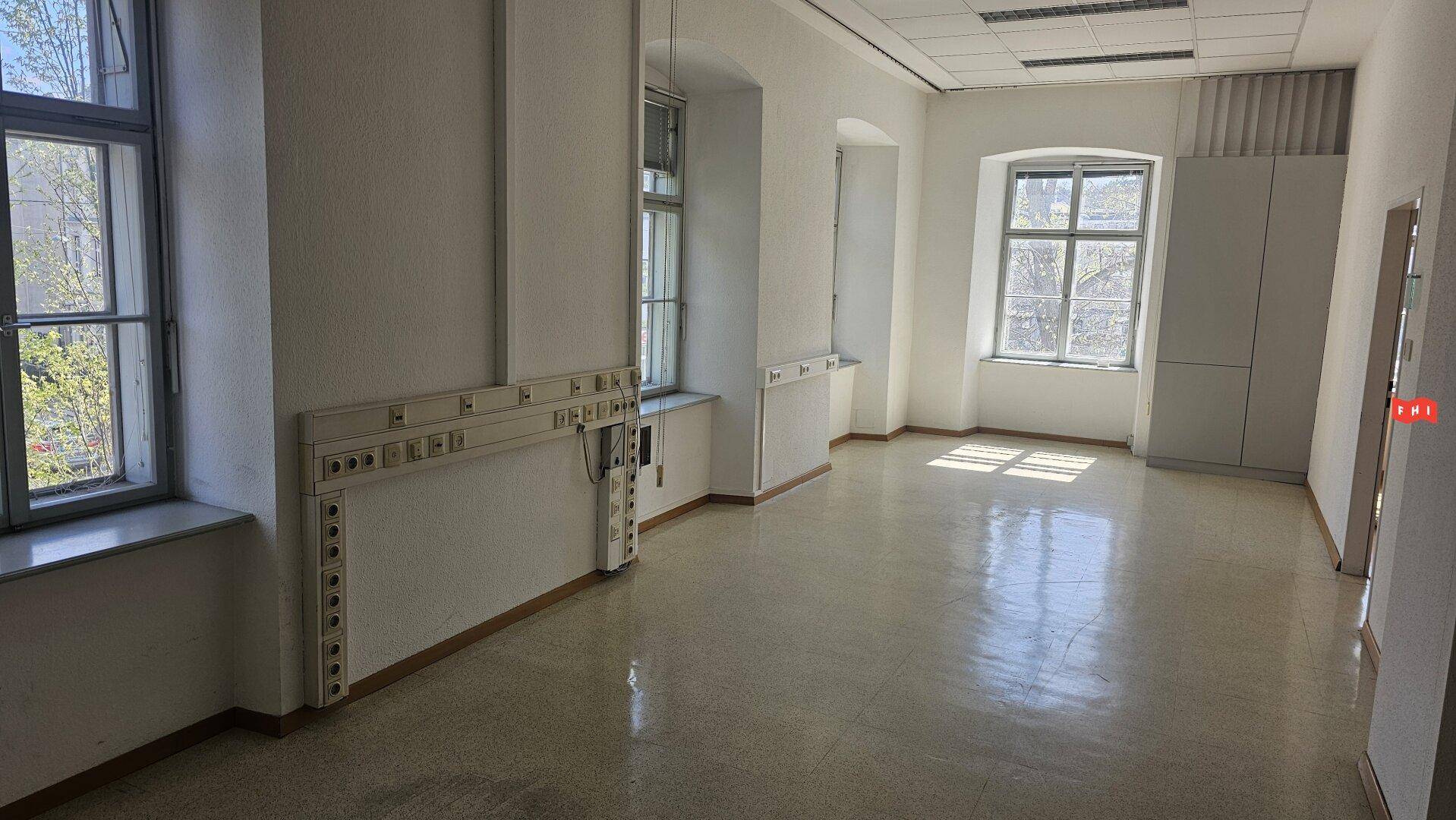 Geräumiger Büroraum mit Terrazzoboden, großen Fenstern und einer Wandinstallation mit Anschlüssen.