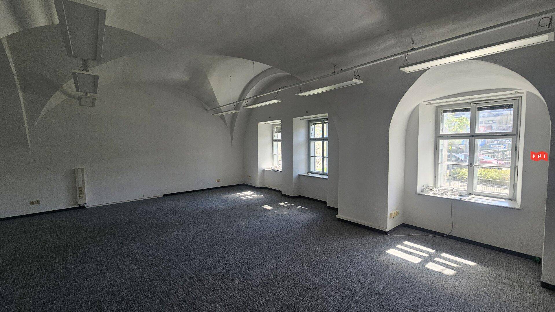 Geräumiger Büroraum mit weißer Gewölbedecke, Teppichboden und großen Fenstern, die viel Licht hereinlassen.