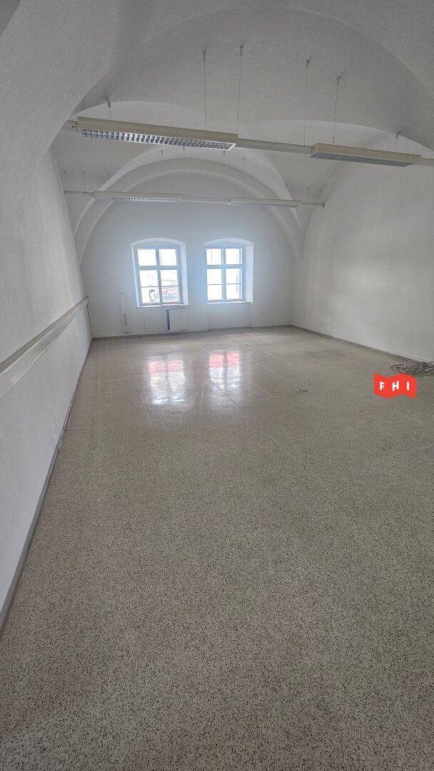 Langer Büroraum mit Gewölbedecke, Terrazzoboden und zwei Fenstern am Ende, die Tageslicht spenden.