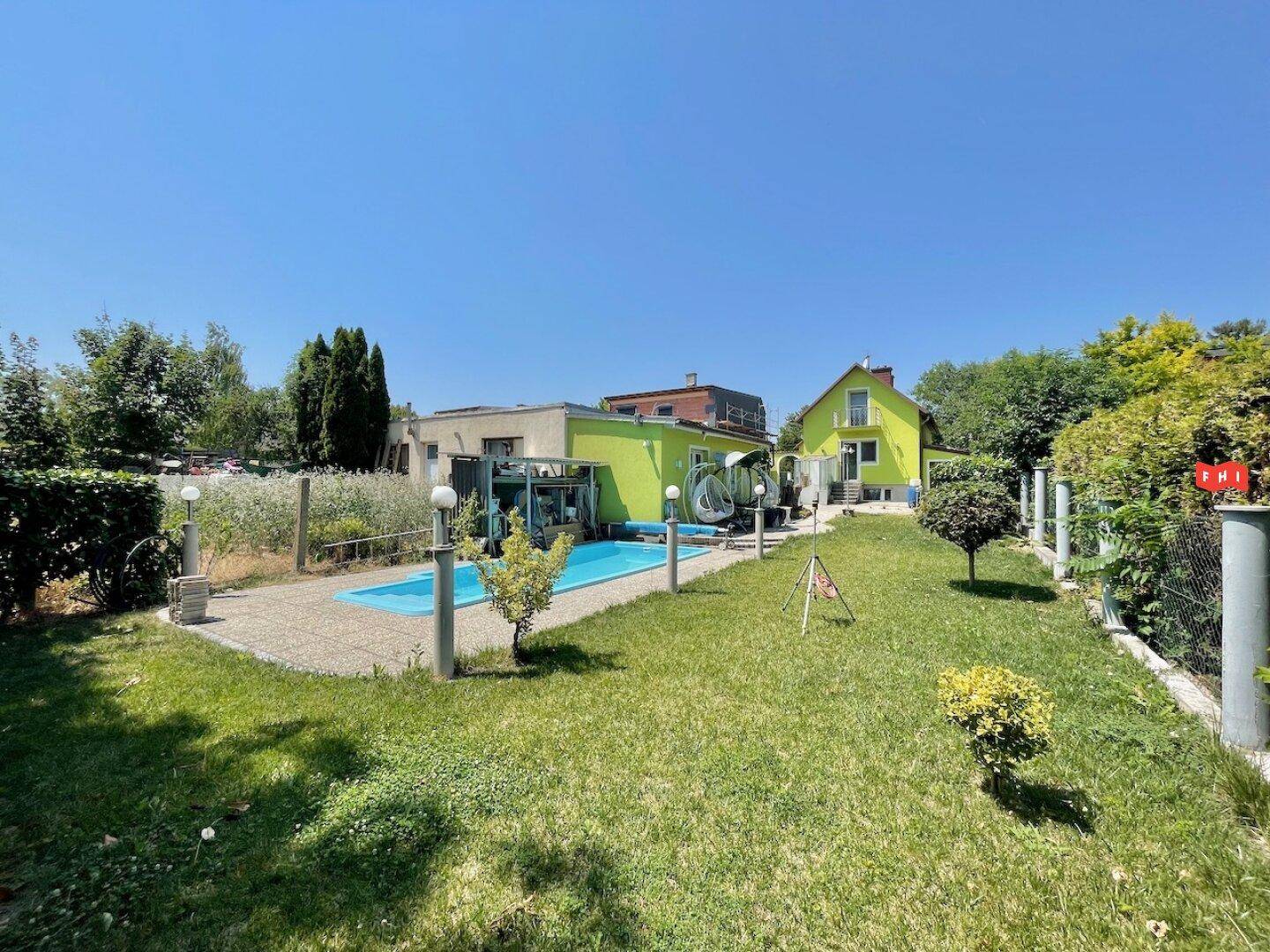 Großer Garten mit Rasenfläche, einem Pool und dem gelbgrünen Haus im Hintergrund unter blauem Himmel.