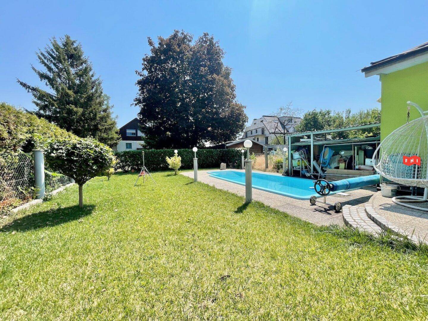 Gepflegter Garten mit blauem Pool, Rasenfläche und hohen Bäumen im Hintergrund.