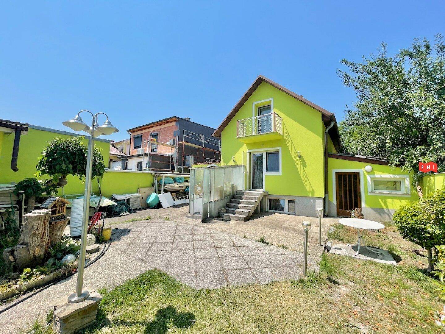 Rückseite des gelbgrünen Hauses mit kleinem Balkon, gepflasterter Terrasse und Zugang zum Garten.