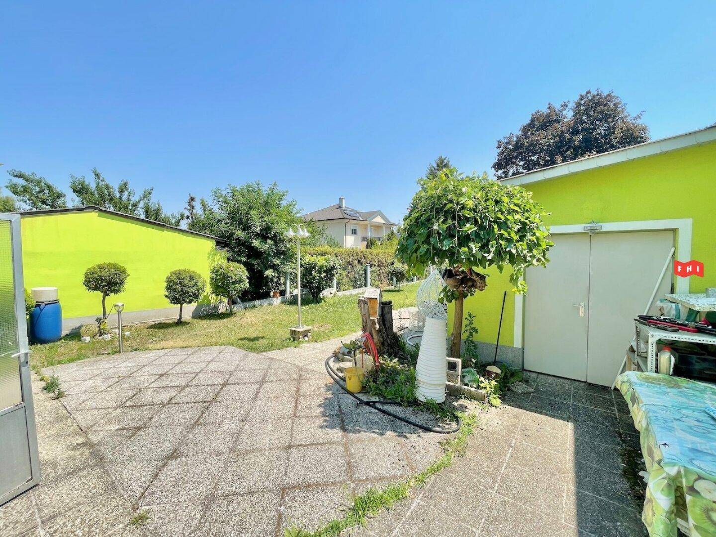 Gepflasterter Hinterhofbereich mit Gartenhaus, Bäumen und Blick auf die Umgebung.