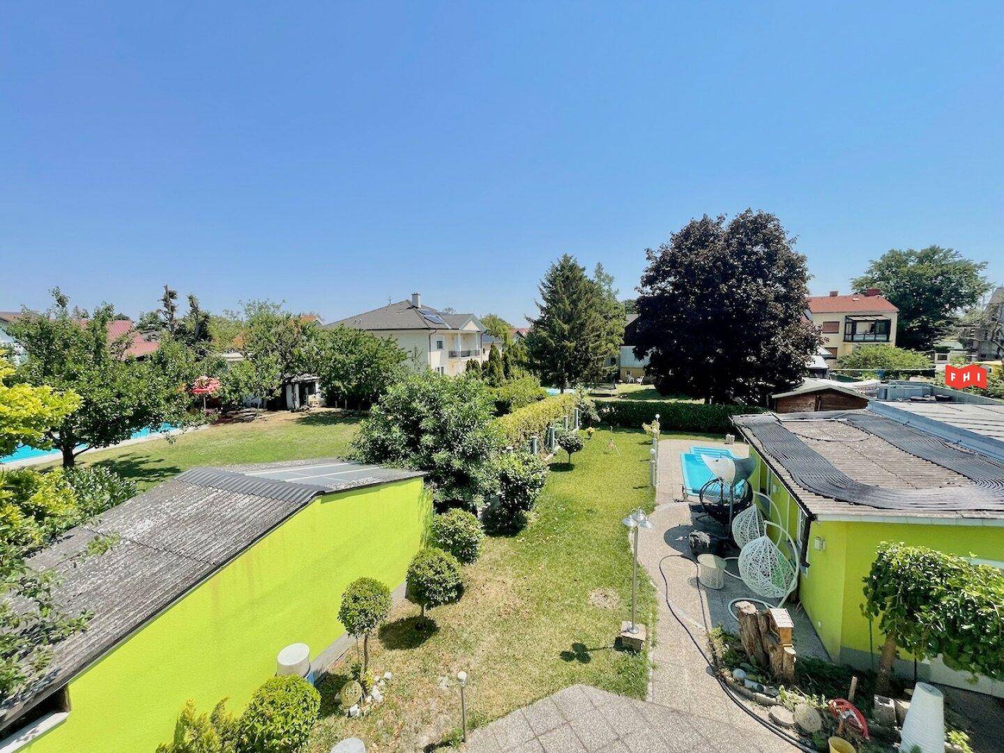 Überblick über den Gartenbereich mit viel Grün, Bäumen und angrenzenden Häusern unter blauem Himmel.