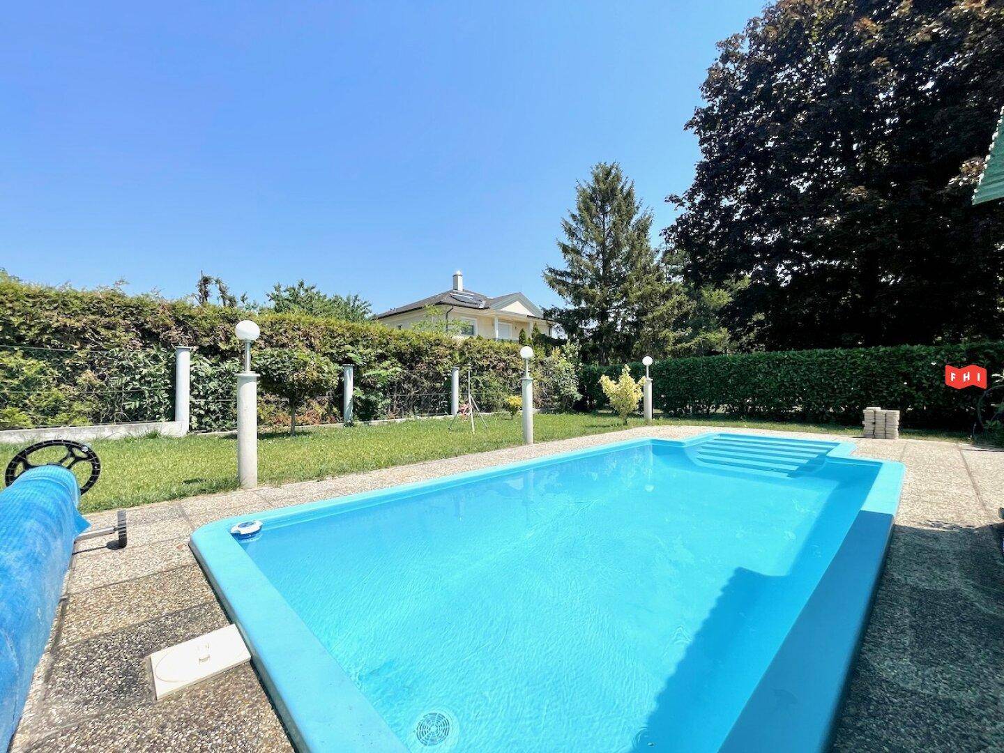 Blauer Swimmingpool mit Treppe, umgeben von Rasen und einer Hecke unter klarem Himmel.