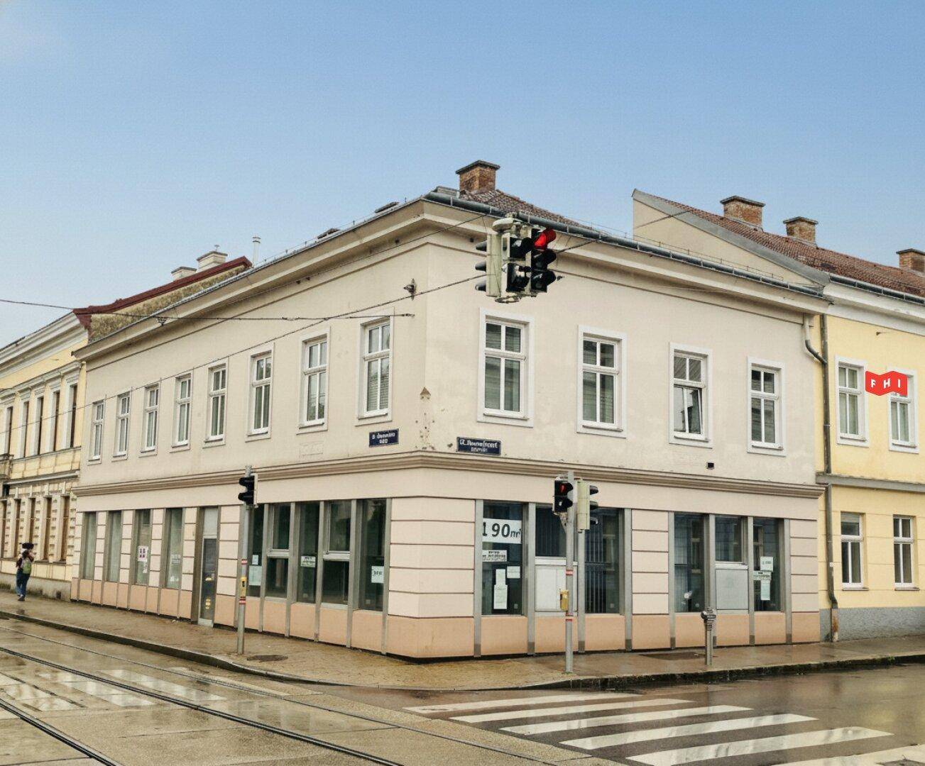 Historisches Eckgebäude mit heller Fassade und großen Schaufenstern im Erdgeschoss an einer Kreuzung.