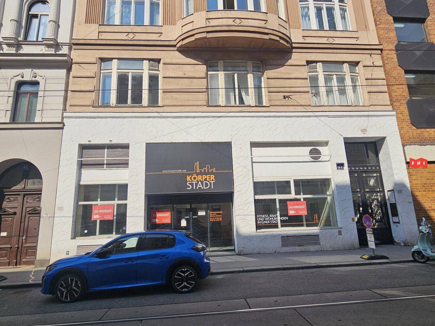 Außenansicht eines mehrstöckigen Gebäudes mit einem Fitnessstudio im Erdgeschoss und einem klassischen Balkon im ersten Stock.