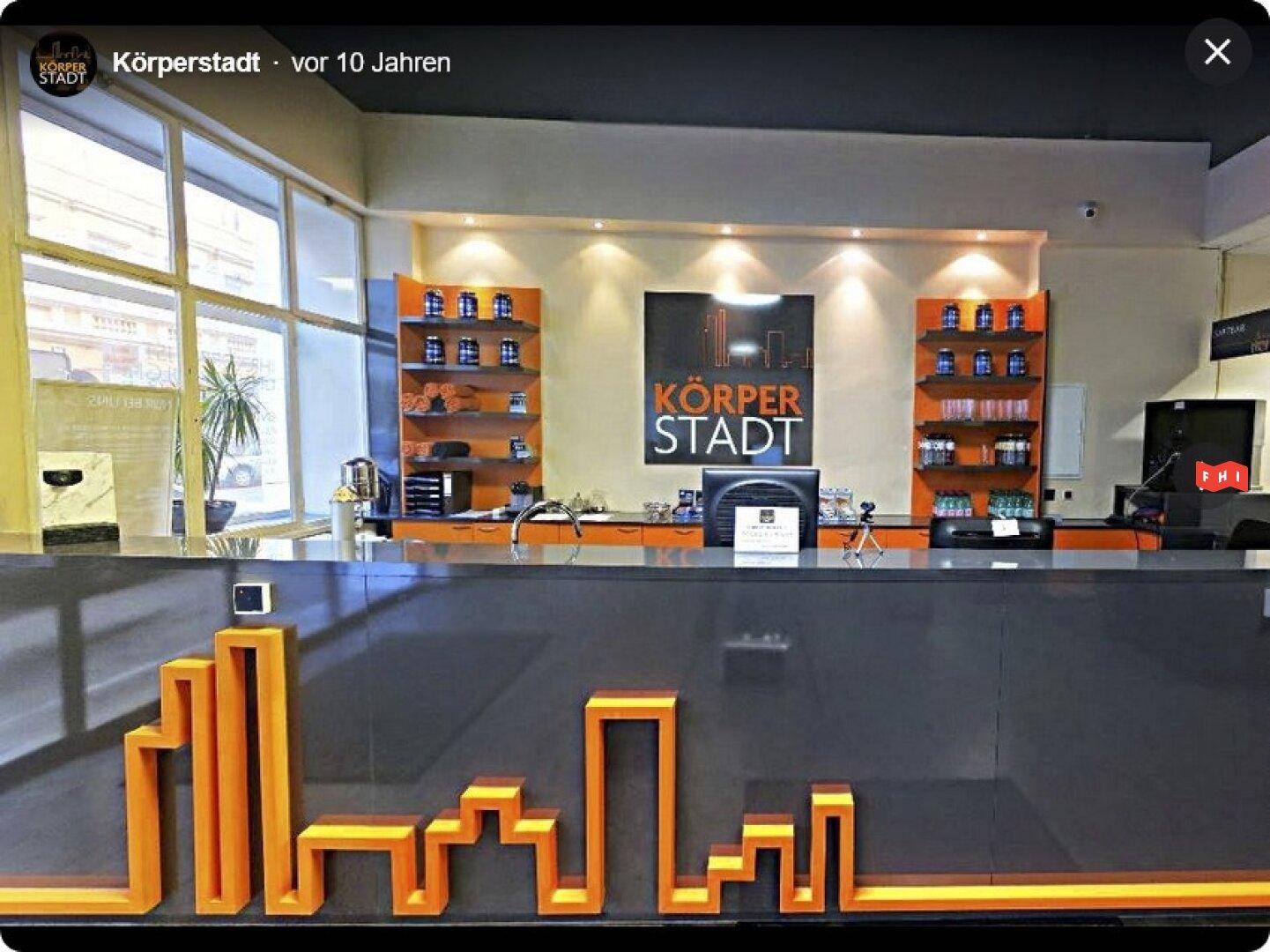 Empfangsbereich eines Fitnessstudios mit einer modernen Theke, Regalen und großen Fenstern, die viel Licht hereinlassen.