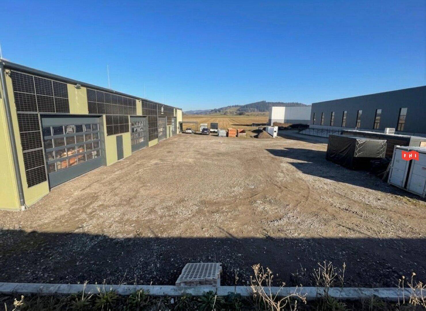 Weitläufiger Schotterplatz vor dem Gewerbegebäude mit weiteren Industriegebäuden im Hintergrund unter klarem Himmel.
