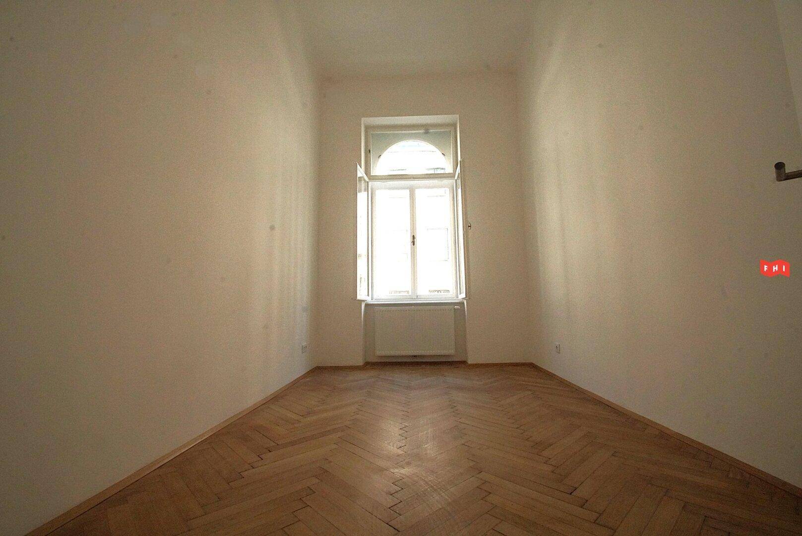 Ein leerer Raum mit Fischgrätparkett und einem hohen Bogenfenster, das viel Licht spendet.