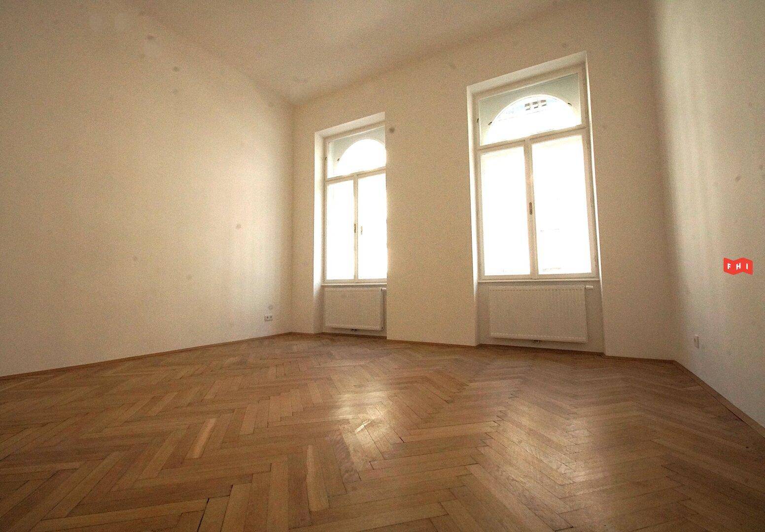 Ein heller Raum mit Fischgrätparkett und zwei hohen Bogenfenstern, die viel Tageslicht bieten.