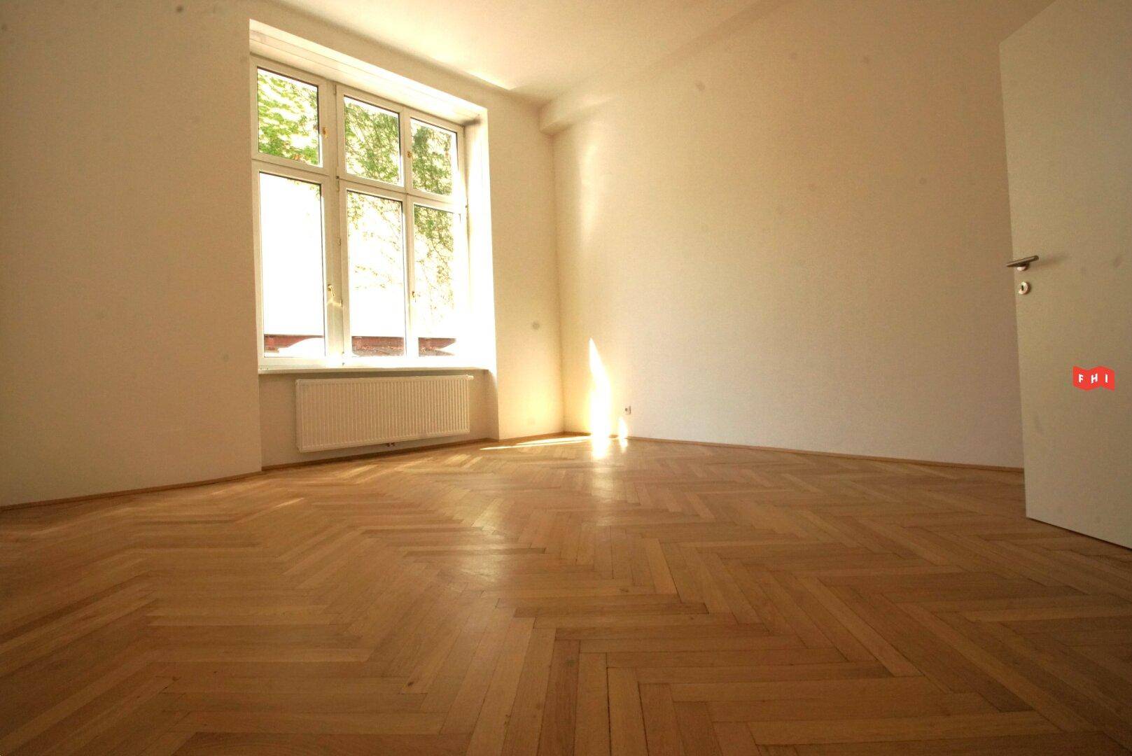 Ein sonniger Raum mit Fischgrätparkett und einem großen Fenster, das viel Tageslicht hereinlässt.