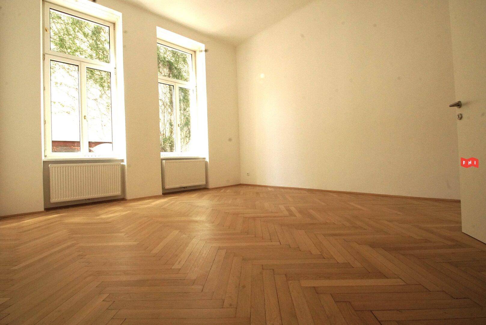 Ein heller Raum mit zwei großen Fenstern, Fischgrätparkett und weißen Wänden, bereit zur Einrichtung.